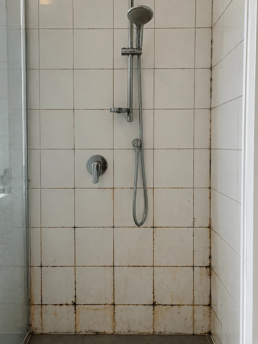 White shower wall tiles with black mould and orange soap-scum staining along the grout lines before tile and grout cleaning